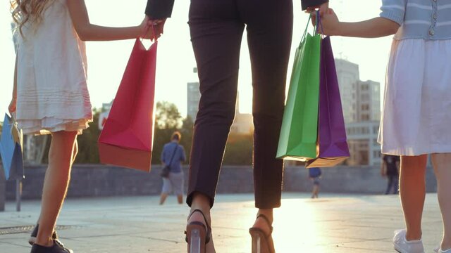 Close up of human legs, two small girls walking down street in stylish shoes, holding mom's hand. Mother in fashionable luxury heels goes with daughters from shopping store, carries in packages gifts