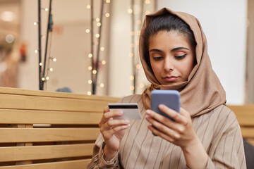 Portrait of young Middle-Eastern woman wearing headscarf making online purchase via smartphone...