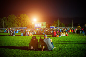 Summer cinema. People and couples watching a movie on the screen of a summer cinema in the evening - obrazy, fototapety, plakaty