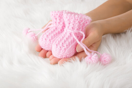 Young woman hands holding pink knitted fluffy baby socks on white fluffy fur blanket background. Closeup.