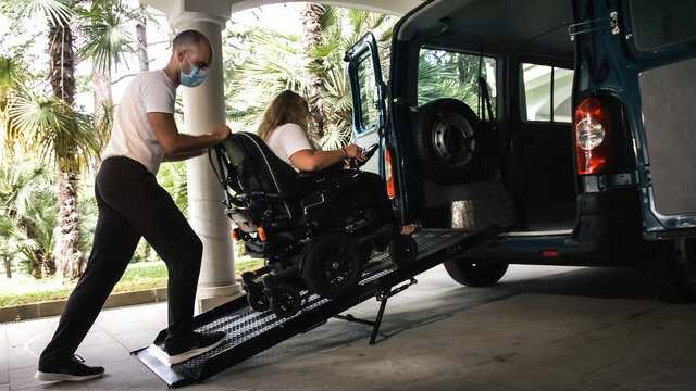 Assisting disabled woman on wheelchair with transport using accessible vehicle with ramp wearing a face mask.
