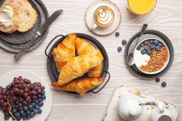 Beautiful morning breakfast with fresh croissants, granola with yoghurt and berries, toasts, coffee cappuccino and fruit on plate. Tasty and healthy breakfast on white wooden table, top view .