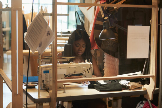Young female designer working at sewing machine behind plexiglass glass in a cover-19 safe workplace.