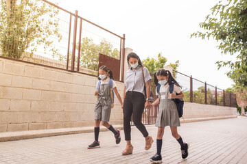 Obraz premium Young mother walking hand in hand with two school girls wearing uniform and face mask before enter school.