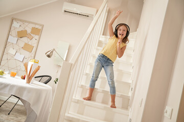 Little singer. Cute and happy caucasian girl wearing headphones standing on stairs and listening music, she is singing and having fun at home