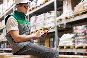 warehouse inventory management. warehouse worker is writing, keeping count, caucasian guy in helmet and uniform is concentrated on work in warehouse