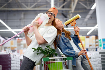happy man and woman enjoy shopping, have fun, sing and dance holding mops, man with bucket on head,...