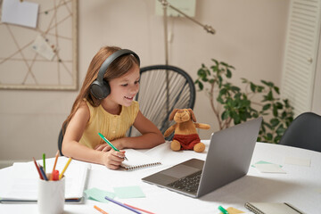 E-learning system. Smart little caucasian girl in headphones sitting at the table, using laptop and...