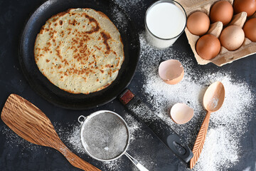 Hot pancake in black pan on black table with flour, milk and eggs