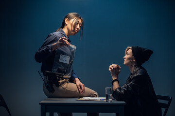 A young asian female investigator sits on a table and shows a package of weapons to a blonde suspect.