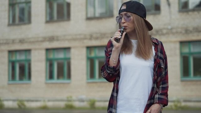 Pretty young hipster woman in black cap vape ecig. Girl vape popular ecig gadget, vaping device.Happy brunette vaper girl with e-cig. Portrait of smoker female model with electronic cigarette