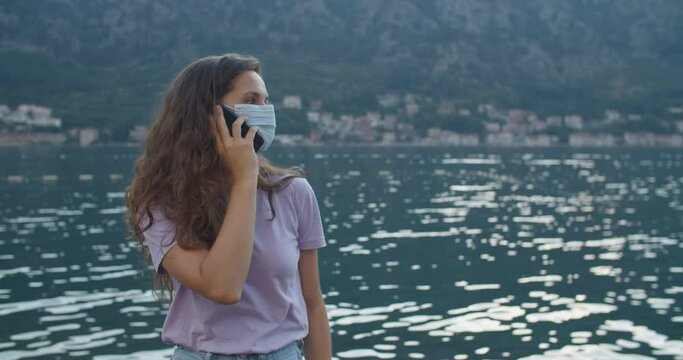 A young woman tourist with curly hair and a protective mask stands on a wooden pier and talks on the phone. Mountain and bay views. Woman portrait close-up. High quality 4k footage