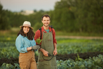 Portrait of two workers in field at vegetable plantation, young woman and man smiling at camera in foreground, copy space