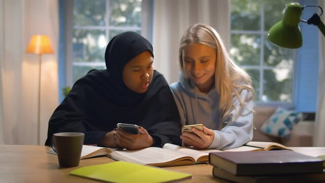 Two multiethnic female students studying together sitting at desk in campus using smartphone
