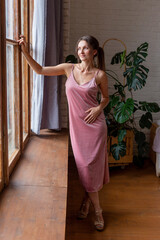 Fototapeta premium Beautiful young woman with dark hair and in a pink dress stands by the window