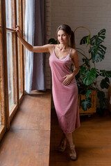 Beautiful young woman with dark hair and in a pink dress stands by the window