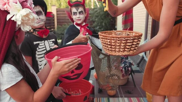 Handheld shot of happy young woman in witch hat holding basket and giving bunch of candy to cute children in halloween costumes