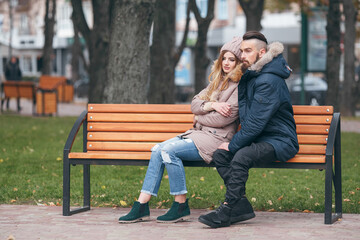 A guy and a girl are resting on a bench in an autumn park. A loving couple in jackets sits on a bench in the main park