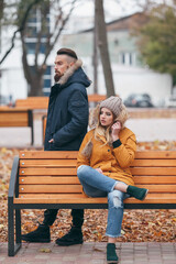 A guy and a girl are resting on a bench in an autumn park. A loving couple in jackets sits on a bench in the main park