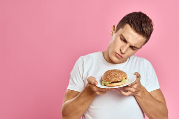 Emotional guy with hamburger on a plate and white t-shirt pink background cropped view of fast food calories