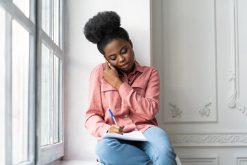 Focused African American biracial woman making notes, clarifies information, talking on phone, sitting on windowsill. Biracial female employee consulting by mobile device, works at home remotely