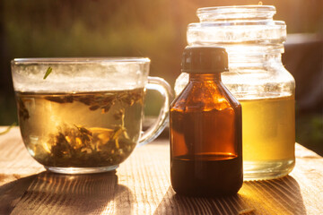 bottle of medicine, liquid spring honey, and transparent cup with brewed St. John's wort on table at sunset. Treatment with herbs. Alternative medicine.