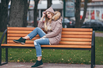 Young woman in autumn clothes sits on a bench in a city park. The woman is dressed in a stylish jacket with fur