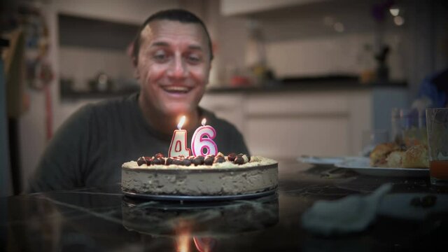 Happy man celebrating his 46th birthday and blowing candles on homemade baked cake, indoor