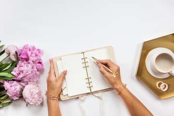 Well-groomed woman hands is writing in diary with stationery supplies set onside and peonies bouquet