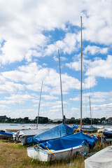 Small sailing or fishing boats on land with masts and covers on in Mudeford Quay, southern England, beautiful blue cloudy sky, many dingies stored on dry shore