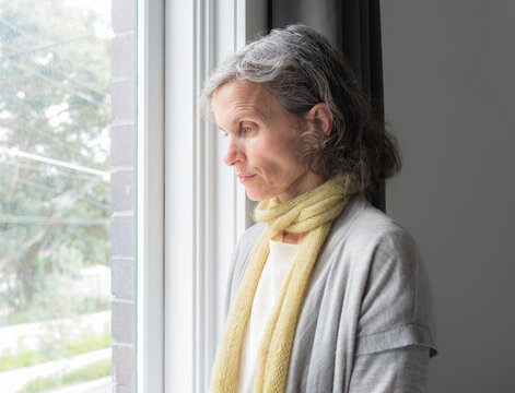 Close up of sad middle aged woman lwith grey hair ooking down next to window (selective focus)