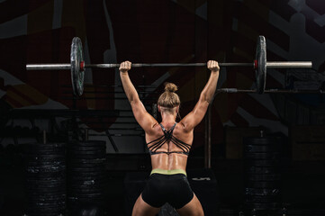 Back view young adult girl doing barbell squats in gym. Woman with muscular body doing lifting...