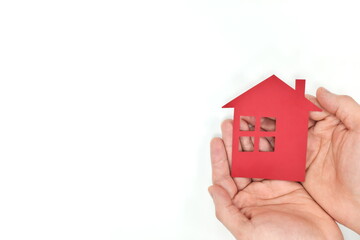 Male cupped hands holding a red house paper cutout in white background. Home insurance, care and protection concept. Top view, flat lay with copy space.