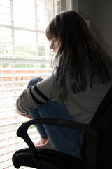 Young woman sitting on a chair in front of a window, holding her legs in front of her up on the chair