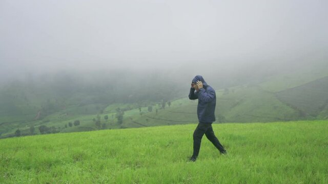 Asian man tourist in blue jacket pulling the hood over his head and walking against strong wind blowing on the mountain in foggy day. Outdoor hiking lifestyle, bad weather and travel vacation concept.