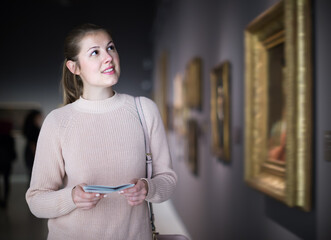 Active girl visitor holding guide book standing near pictures in museum of arts