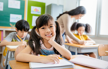 Portrait of smile little pupil writing at desk in classroom at the elementary school. Student girl doing test in primary school. Children writing notes in classroom. Education knowledge concept