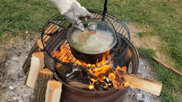 Checking cast iron pot of vegetable broth being prepared for fish pot while boiling over fire pit.