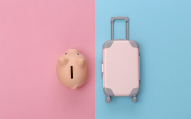 Travel concept. Mini plastic travel suitcase and piggy bank on pink blue pastel background. Minimal style. Top view, flat lay