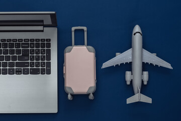 Flat lay vacation holiday and travel planing concept. Laptop and mini plastic travel suitcase, air plane on classic blue background. Top view