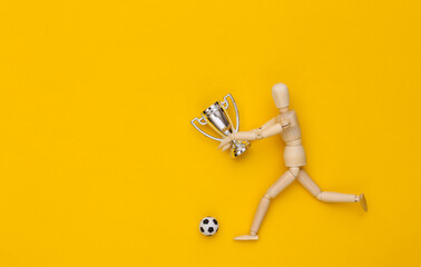 Wooden puppet runs with soccer ball and championship cup on yellow background