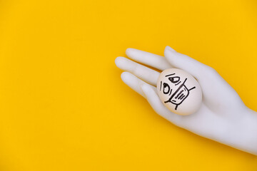 White mannequin hand holds egg face in medical mask on yellow background. Covid-19 pandemic. Top view