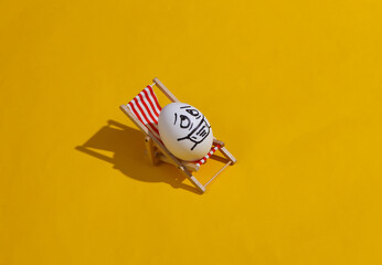 Chicken egg with hand drawn happy face on beach deck chair. Yellow bright background. Summer rest,...