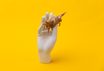 White mannequin hand holds seashell on yellow background