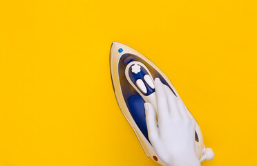 White mannequin hand holds iron on yellow background. Top view