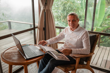 Happy smiling journalist stylish guy read a book  in a workplace in loft styled coworking, well dressed, sitting near window with view of garden lok at the camera