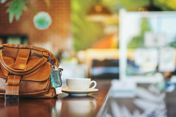 Mini alcohol gel bottle to kill Corona Virus(Covid-19) hang on a brown leather shoulder bag with hot coffee on table in cafe.New normal lifestyle.Health care concept.Selective focus on alcohol gel
