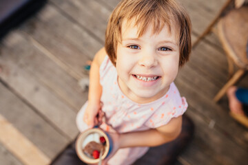 Top view of cute funny little girl with brown hair holding cup with dessert or ice cram. Portrait of cheerful little girl looking up in summer pink dress. - Powered by Adobe