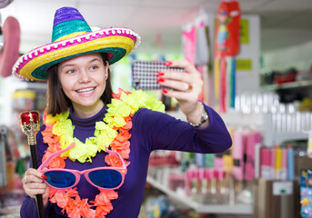 Comically dressed happy girl making funny selfies photo in festive accessories shop