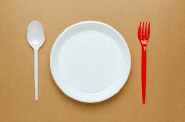 Top view of plastic tableware, white spoon and plate, red fork on the brown surface